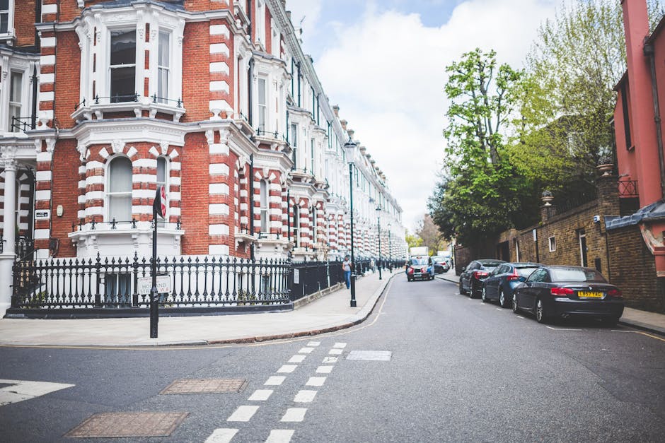 A view of a residential street in Earls Court with Victorian-style terraced houses featuring red brick facades, white decorative window frames, and ornate cornices. The houses are set behind black wrought-iron fences, with some displaying small garden areas. Parked cars line the narrow street, including a black vehicle closest to the foreground and various other cars further down. A large tree with green foliage extends over part of the street, providing shade. The sky is partly cloudy with patches of blue visible. In the distance, a white van is parked near the curb, likely involved in a home relocation or furniture transport process. The street appears calm, with a few pedestrians visible along the pavement. This scene reflects a typical setting where professional removals services, such as those offered by [COMPANY_NAME], may operate to assist with packing, loading, and transporting household items during a house move in the Earls Court area.