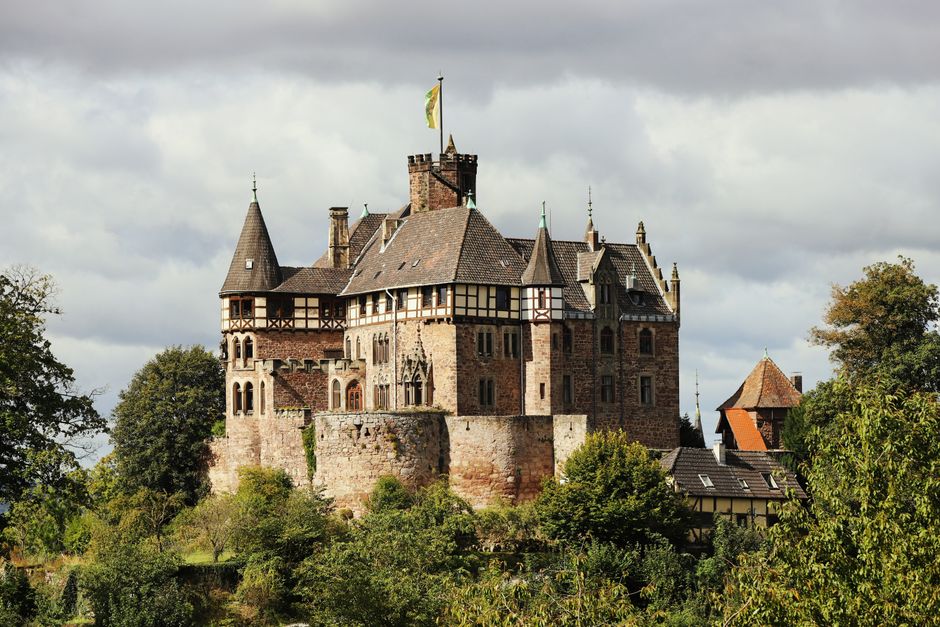 A historic stone castle with multiple towers and turrets, featuring conical roofs, is situated atop a hill surrounded by lush green trees. The castle showcases medieval architectural elements including arched windows, battlements, and a flag flying from the highest tower. The foreground includes a variety of foliage and trees that partially obscure the lower part of the structure, while the sky above is overcast with grey clouds. This scene exemplifies traditional European castle architecture and is depicted in a natural outdoor setting. This image is relevant for house removals and relocation services, as it highlights the importance of careful handling and logistics when moving valuable or delicate items. The detailed visual scene supports the concept of home relocation, furniture transport, and packing and moving processes that [COMPANY_NAME] offers, often working in historic or residential properties similar to those found near Nevern Place in Earls Court.