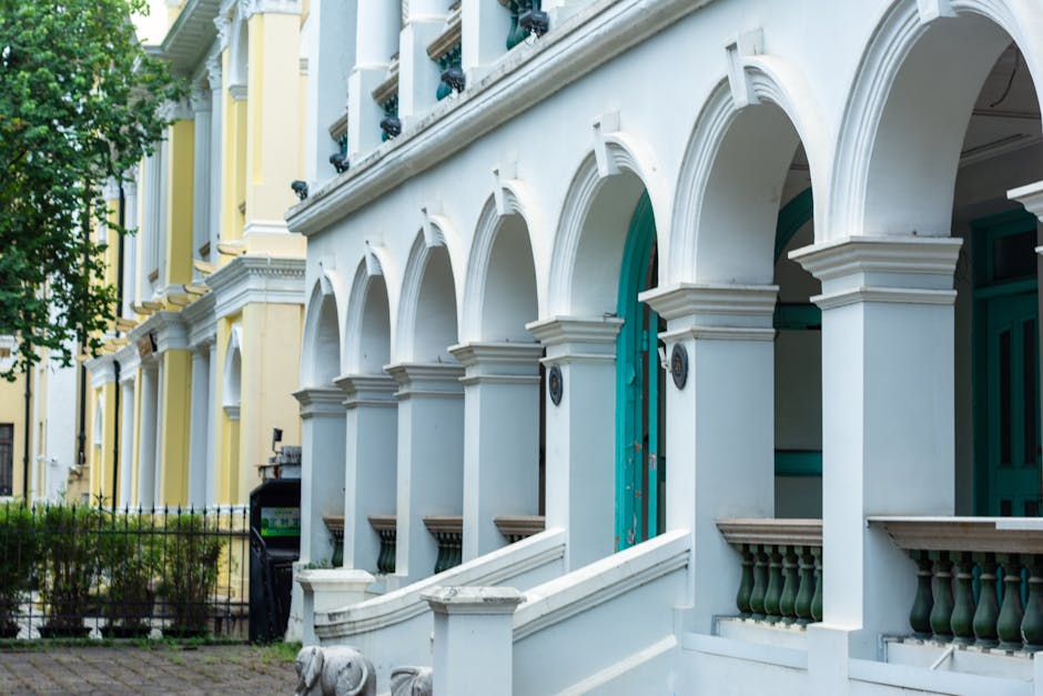 A view of a residential street in Earls Court with Victorian-style terraced houses featuring red brick facades, white decorative window frames, and ornate cornices. The houses are set behind black wrought-iron fences, with some displaying small garden areas. Parked cars line the narrow street, including a black vehicle closest to the foreground and various other cars further down. A large tree with green foliage extends over part of the street, providing shade. The sky is partly cloudy with patches of blue visible. In the distance, a white van is parked near the curb, likely involved in a home relocation or furniture transport process. The street appears calm, with a few pedestrians visible along the pavement. This scene reflects a typical setting where professional removals services, such as those offered by [COMPANY_NAME], may operate to assist with packing, loading, and transporting household items during a house move in the Earls Court area.