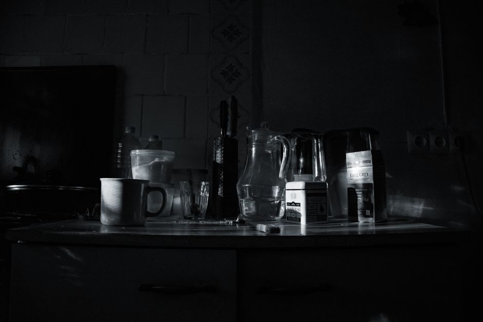 A dimly lit kitchen countertop inside a home, featuring various items associated with packing and house removal processes. Visible objects include a white ceramic mug, a small glass, a plastic jug, an
