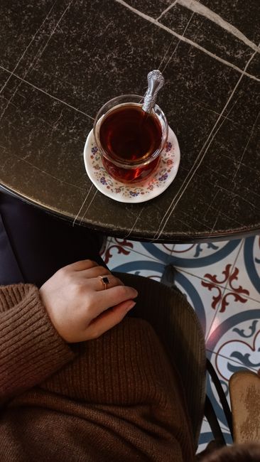 A top-down view of a dark, polished marble or granite table surface on which a transparent glass of hot tea with a metal spoon resting inside is placed on a small floral-patterned saucer. The glass is filled with a dark amber liquid. In the lower left corner, a person's hand and forearm are visible, resting on a brown knitted sleeve, with fingers relaxed over a dark coloured chair. Beneath the table, a patterned floor displays a combination of blue, white, and red decorative tiles. The scene suggests a cozy indoor environment, possibly a cafe or kitchen area, where home relocation or packing activities might be taking place, with this image representing a moment of pause amid moving preparations, as referenced by the context of house removals and furniture transport.