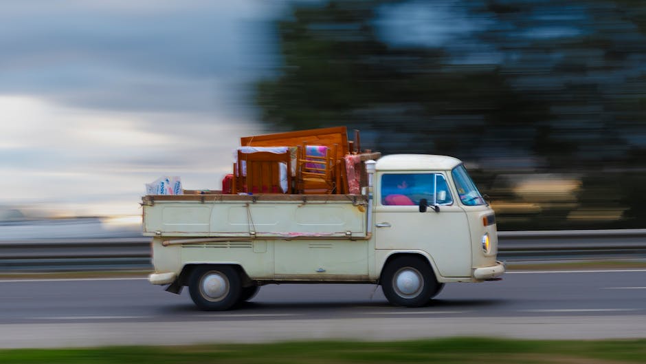 A vintage cream-coloured van with a compact, rounded design is shown in motion on a roadway, loaded with various household items for house relocation, including wooden chairs, a small table, cardboard boxes, and plastic-wrapped packages, all stacked in the open cargo area secured with ropes. The van is passing a metal guardrail, with a blurred background of trees and sky indicating movement. The items inside appear to be part of a packing and moving process, likely transported to or from a residence, and the scene captures a typical furniture transport scenario associated with house removals, as could be relevant to services like those offered by manwithvanearlscourt.org.uk.